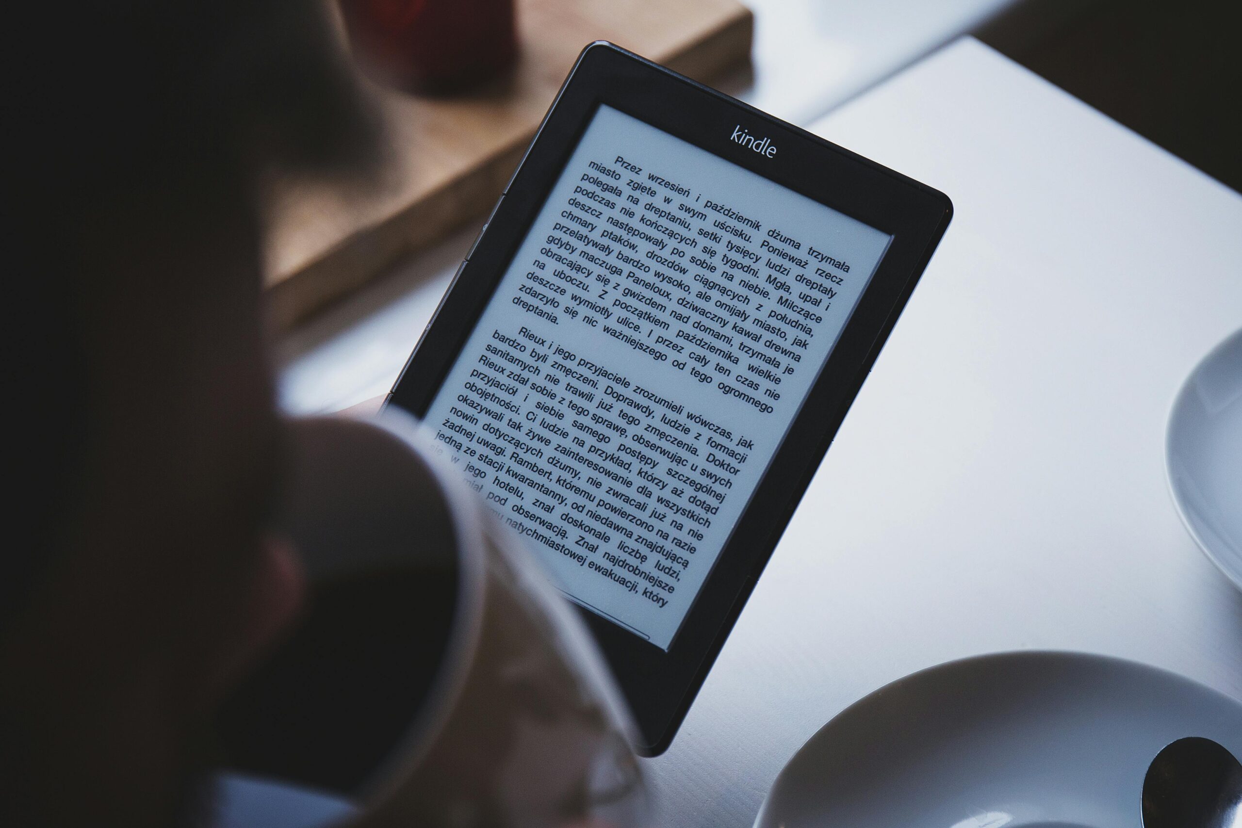 A person reads on a Kindle e-reader while holding a cup of coffee, indoors.